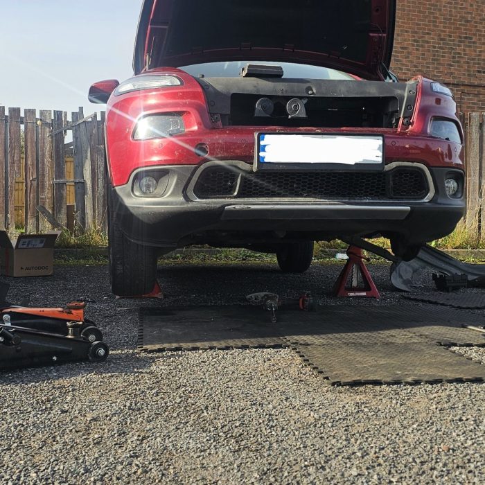 A red car (SUV/crossover) with the hood raised and the front end lifted on jacks and jack stands, undergoing repair or service outdoors on a gravel surface. Tools, jacks, parts (including a detached wheel and underbody shield), and a white van are visible nearby.